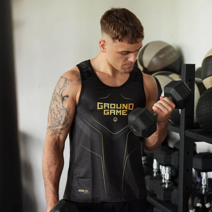 Mężczyzna ćwiczy bicepsy ze sztangielką na siłowni w czarnym tank topie Ground Game Gold 2.0